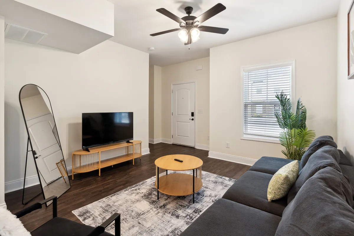 A spacious living area features a plush, modern sofa set facing a television and a large decorative mirror. A coffee table is centered in the room, and a leafy plant adds a touch of greenery. The entire space is bathed in natural light from large windows that are not directly visible but inferred by the brightness and reflections.