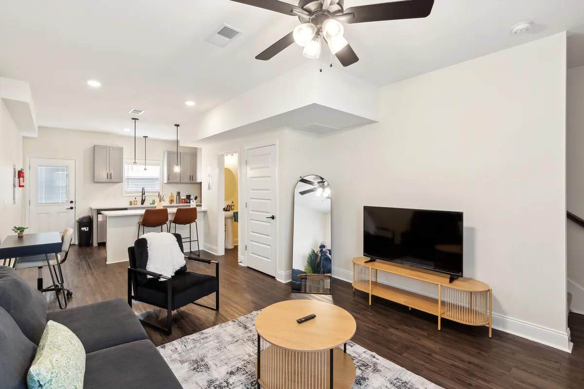 The image showcases a stylish open-plan living area with a plush sofa, sleek coffee table, large wall-mounted TV, and a modern kitchen island with bar stools. The space is bright, inviting, and perfect for relaxing, entertaining, or enjoying a cozy night in.