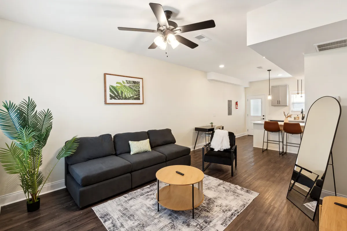 A modern living space with a comfortable sofa, a sleek coffee table, and a potted plant, accented by wall art and a view of adjacent bar stools. The open layout and stylish décor create an inviting atmosphere suited for both quiet evenings and casual entertaining.