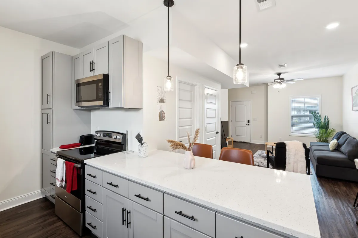 An inviting kitchen with a central island, bar stools, and modern stainless steel appliances. The open-plan layout also connects to a living area, emphasizing the social and communal aspects of cooking and dining at home.