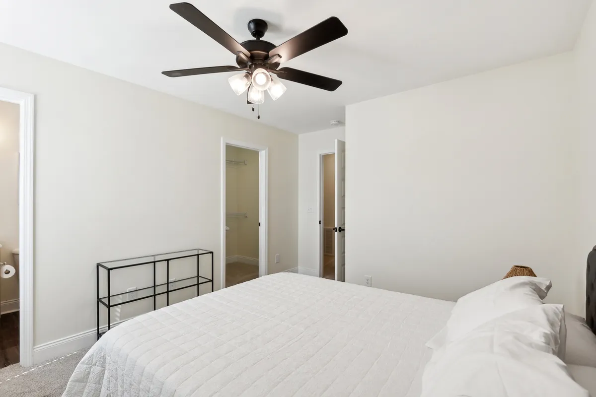 A tranquil bedroom scene featuring a neatly made bed, a nightstand, ceiling fan, and open doorways that hint at the interconnected living spaces beyond. The room is softly lit, creating a calm and inviting atmosphere.