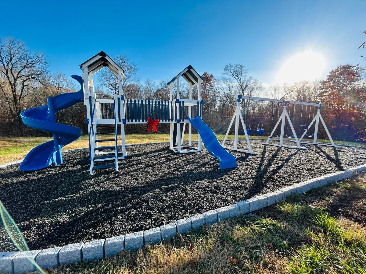 A lively exterior scene centered on a colorful playground structure featuring a blue slide and swings. Children are actively playing while families gather and enjoy picnics in the surrounding grassy area, evoking a joyful, family-friendly atmosphere. The setting is perfect for birthday parties, playdates, and casual outdoor fun.